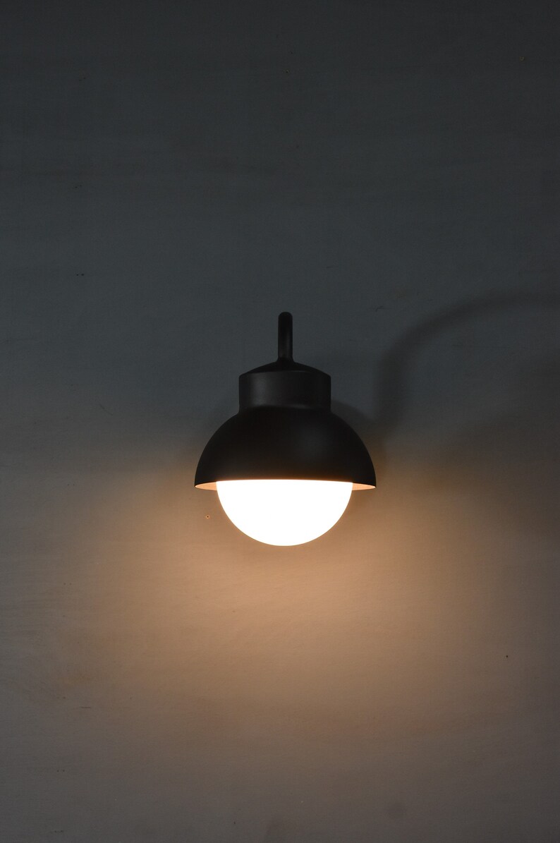 Op de afbeelding: Een zwarte wandlamp met een ronde, witte bolvormige kap. Het licht is aan en verlicht de omgeving.