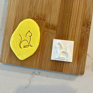 May include: A white plastic cookie cutter in the shape of a cat sitting down. The cutter is on a wooden cutting board with a yellow piece of modeling clay that has been pressed with the cutter.