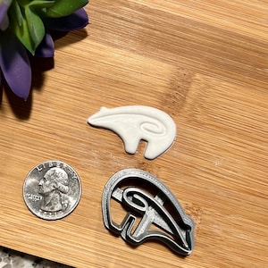 May include: Two cookie cutters, one silver and one white, shaped like a Native American symbol. The silver cutter is on a wooden surface.