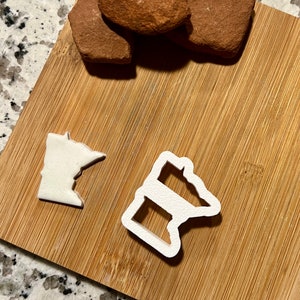May include: Two white plastic cookie cutters shaped like the state of Minnesota. One cutter is a solid outline of the state, and the other has a horizontal stripe across the middle.
