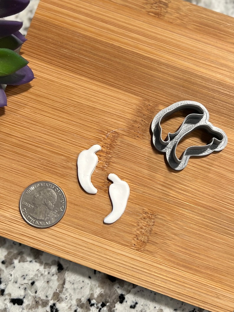 May include: Two white plastic chili pepper cookie cutters and a silver metal chili pepper cookie cutter on a wooden surface.