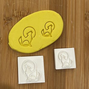 May include: Two white plastic cookie cutters with a wolf howling at the moon design. The cutters are on a wooden surface. One cutter is on a yellow silicone sheet with the same design.