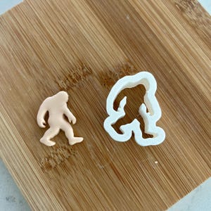 May include: Two cookie cutters in the shape of Bigfoot. The larger cutter is white and the smaller cutter is a light brown color.