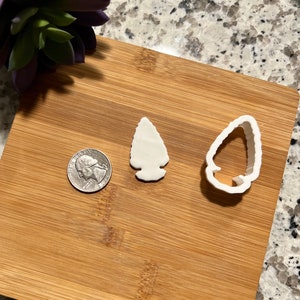 May include: Two white plastic cookie cutters shaped like arrowheads. The cutters are on a wooden surface next to a US quarter.