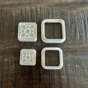 May include: Four white plastic cookie cutters, two with a floral design and two square shaped.