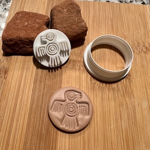 May include: A white plastic cookie cutter and a pressed clay circle with a bird design. The cutter and clay are on a wooden cutting board with two reddish-brown rocks. The bird design has a circular body and outstretched wings.