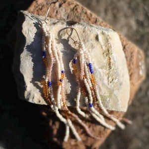 May include: Beaded dangle earrings with a cream, blue, and gold color scheme. The earrings feature multiple strands of small beads, with gold-colored ear wires. The earrings are displayed on a light-colored stone surface.