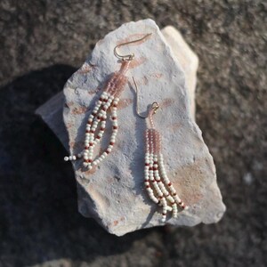 May include: A pair of beaded earrings with a gold-colored hook. The earrings feature a light pink top section and white, red, and pink beads that hang down in a fringe style. The earrings are displayed on a light-colored rock.