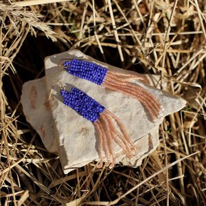 May include: A pair of handmade beaded earrings. The earrings feature a vibrant blue beaded top section that transitions into a cascade of peach-colored beads. The earrings are displayed on a light-colored stone, set against a backdrop of dry grass.