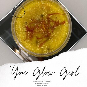 May include: A glass jar filled with a yellow body scrub with orange flower petals. The text "You Glow Girl" is written in black cursive script. The text "CALENDULA, TUMERIC, & MANUKA HONEY BODY SCRUB" is written in black below the cursive text.