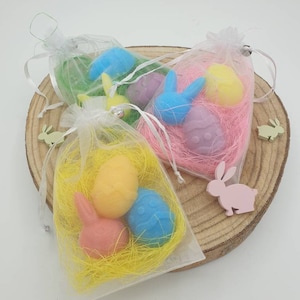 Könnte beinhalten: Drei kleine Organzasäckchen gefüllt mit pastellfarbenen Ostereiern und Hasenformen. Die Säckchen sind mit gelbem, rosa und grünem Schredderpapier gefüllt.