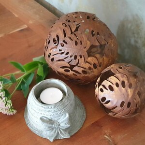 May include: Three decorative candle holders, two are round and made of metal with a rust-colored finish and cut-out leaf designs. The third is a smaller, gray concrete holder shaped like a hat with a bow.