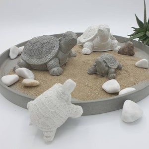 Könnte beinhalten: Ein Set aus vier kleinen Beton-Schildkrötenfiguren in verschiedenen Posen, auf einem runden grauen Tablett mit Sand und weißen Steinen.