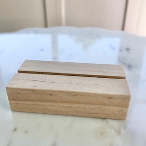May include: Two small, rectangular wooden blocks with a natural finish. The blocks are light brown and have a slot cut into the top. The blocks are likely used as a display stand or holder for signs or photos.