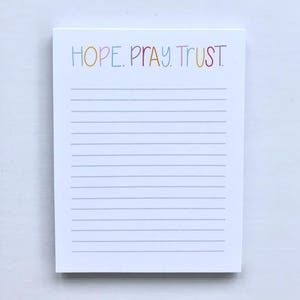 May include: A white notepad with horizontal gray lines. The words "HOPE. PRAY. TRUST." are written across the top in colorful, handwritten-style letters. The notepad is rectangular and sits on a white surface.