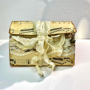 May include: A small box covered in sheet music with a white lace trim and a white ribbon tied around it. The box has a small metal charm hanging from it.