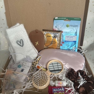 May include: A gift box containing self-care products. It features a white fluffy sock with a heart, a brown luggage tag, eye masks, a Garnier sheet mask, a pink satin eye mask, a small mirror, a hairbrush, sweets, and a scrunchie.