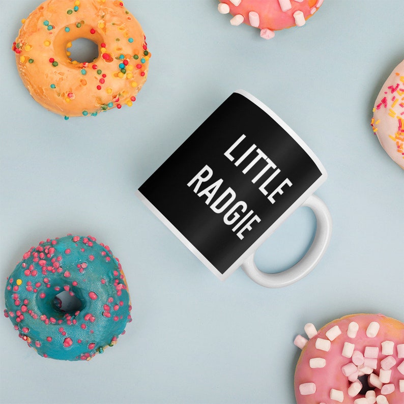 May include: A black coffee mug with white text that reads "LITTLE RADGIE". The mug is surrounded by colorful donuts with sprinkles and marshmallows.