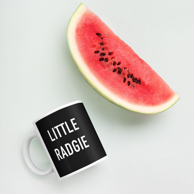 May include: A white ceramic mug with a black handle and a black background. The text "LITTLE RADGIE" is printed in white on the mug. A slice of watermelon is in the background.