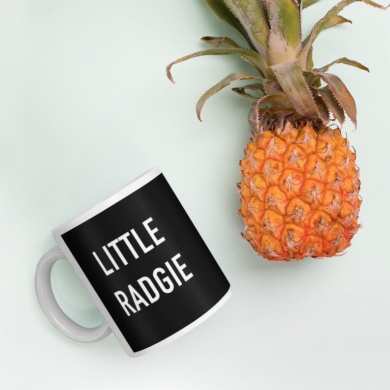May include: A white ceramic mug with a black handle and a black design that says "LITTLE RADGIE" in white text. The mug is sitting on a light green surface next to a small pineapple.