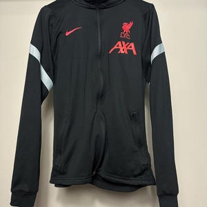Könnte beinhalten: Schwarze Jacke mit Reißverschluss, rotem Nike-Logo und rotem Emblem mit Vogel und den Texten "LFC" und "AXA". Die Jacke hat einen hohen Kragen, lange Ärmel und silberne Streifen an den Oberarmen.