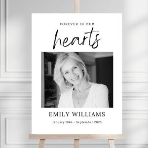 May include: A memorial sign on a wooden easel. The sign features a black and white photograph of a woman, with the text "FOREVER IN OUR hearts" above the photo. Below the photo is the name "EMILY WILLIAMS" and the dates "January 1946 - September 2025."