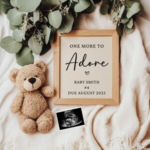 Könnte beinhalten: Ein gerahmtes Schild mit dem Text "One More To Adore" und einem Herzsymbol. Unter dem Text steht "Baby Smith #4 Due August 2025". Ein Teddybär und ein Ultraschallbild liegen auf einer weißen Decke.