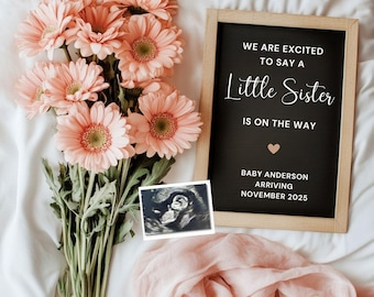 May include: A pregnancy announcement featuring a black chalkboard sign with the text "WE ARE EXCITED TO SAY A Little Sister IS ON THE WAY." The sign is framed in light wood, with peach gerbera daisies, a sonogram, and a pink scarf.