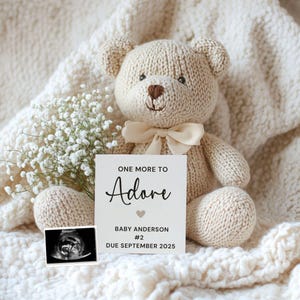 Puede incluir: Una tarjeta blanca con texto negro que dice "One More to Adore Baby Anderson #2 Due September 2025". Un oso de peluche blanco tejido se encuentra junto a la tarjeta. Una pequeña imagen de ultrasonido está en la esquina inferior izquierda de la tarjeta.