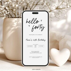May include: A smartphone displays a birthday invitation with the words "hello forty" in elegant script. The invitation includes details for Anna's 40th birthday, with a heart symbol. A white heart-shaped decoration sits beside the phone.