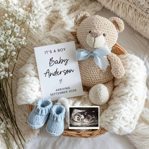 May include: A baby announcement with a knitted teddy bear, blue booties, and an ultrasound image. The announcement card reads "IT'S A BOY Baby Anderson ARRIVING SEPTEMBER 2026". The items are arranged in a woven basket on a white, textured blanket.