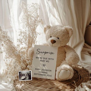 Könnte beinhalten: Ein cremefarbener Teddybär hält eine Karte mit der Aufschrift "Surprise, Wir erwarten Baby #2, Baby Johnson kommt im September 2025." Ein Ultraschallbild ist zu sehen. Der Bär sitzt auf einem geflochtenen Korb.