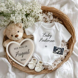 Puede incluir: Un arreglo de anuncio de bebé en una cesta de mimbre. Incluye un body blanco con "Baby Anderson" impreso, un osito de peluche, un letrero en forma de corazón que dice "See You In August 2026", patucos y una imagen de ultrasonido.