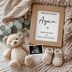 Könnte beinhalten: Ein gerahmtes Schild mit dem Text "Here we grow again" mit einem Herzsymbol, "Baby Wilson #2" und "Due August 2025". Das Schild ist umgeben von einer Strickdecke, einem Teddybär, einem Paar Strickschühchen und einem Ultraschallbild.