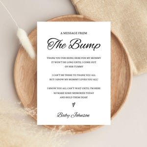 May include: A white card with the text "A MESSAGE FROM The Bump" and a message from the baby. The card is on a wooden plate with a beige linen cloth in the background. The baby's name is "Baby Johnson".