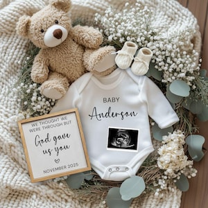 May include: A baby announcement featuring a white onesie with the name "Anderson" and a sonogram image. A teddy bear, baby booties, and a framed sign that reads "We thought we were through but God gave us you" are also present. The announcement is set against a backdrop of flowers.
