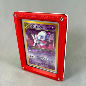 May include: A rectangular red plastic display frame holding a Japanese trading card featuring a purple Espeon character. The frame has four silver metal screws at the corners, securing the card behind a clear protective layer for display purposes.