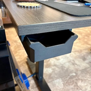 Può includere: Un cassetto portaoggetti nero opaco montato sotto una scrivania con venature del legno. Il cassetto presenta un design a giorno con un bordo anteriore incassato, offrendo una soluzione di archiviazione compatta fissata al telaio.