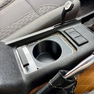 May include: A black plastic cup holder insert designed for a vehicle center console. The circular holder features a smooth interior and a rectangular base plate with a small black lever on the left side, positioned near a gear shifter and seat.