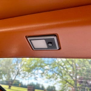Könnte beinhalten: Eine rechteckige Innenraumleuchte für Fahrzeuge, die in eine orangefarbene, strukturierte Verkleidung eingelassen ist. Die Leuchte verfügt über eine weiße rechteckige Linse neben einem kleinen schwarzen runden Druckknopf in einem silbernen Rahmen.
