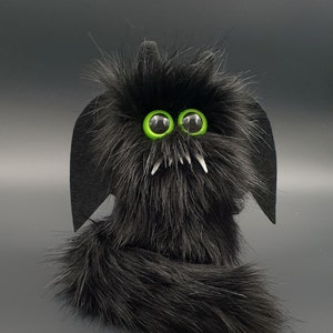 Op de afbeelding: Een zwart pluche speelgoed met grote groene ogen en tanden. Het speelgoed heeft een lange staart en vleermuisvleugels.