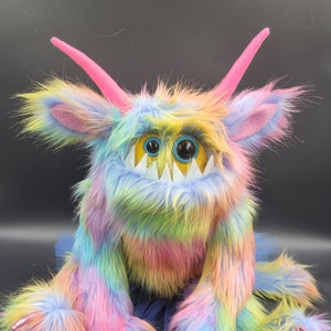 May include: A vibrant, plush monster toy with rainbow-coloured fur, pink horns, and large blue eyes. The creature has sharp white teeth, pink claws, and a blue tutu. The monster's fur is a mix of pink, yellow, blue, and green.