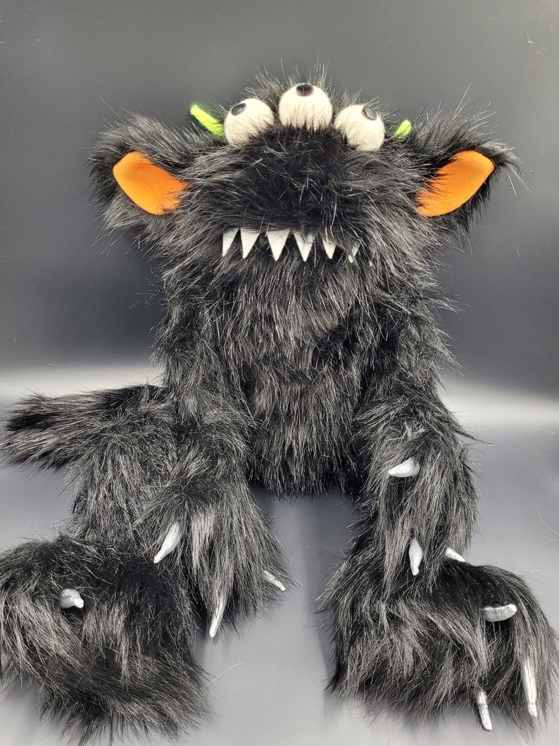 May include: A black furry monster puppet with three white eyes, orange ears, and sharp white claws. The monster has a wide, toothy grin.