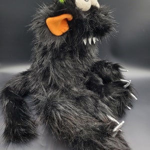 May include: Black furry monster puppet with a green horn, orange ear, and large white eyes. The puppet has sharp silver claws and a toothy grin.