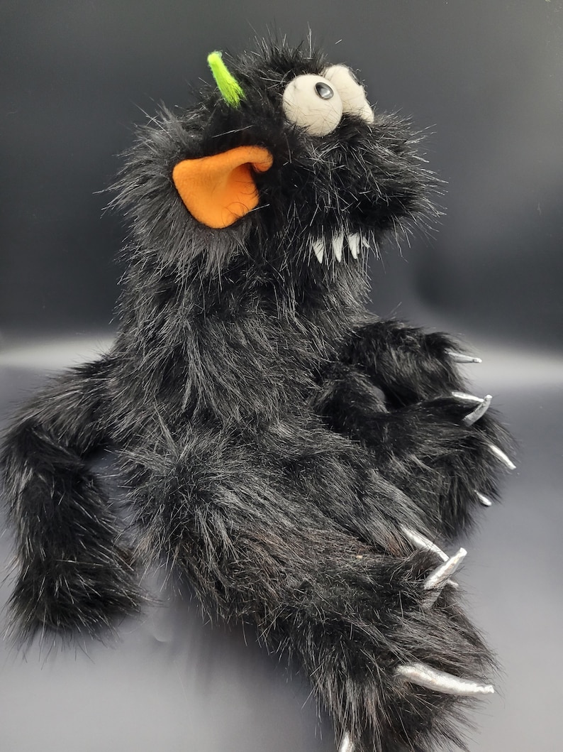 May include: Black furry monster puppet with a green horn, orange ear, and large white eyes. The puppet has sharp silver claws and a toothy grin.