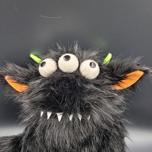 May include: A black furry monster toy with three large white eyes, orange ears, and a toothy grin. The monster has green antennas on top of its head.