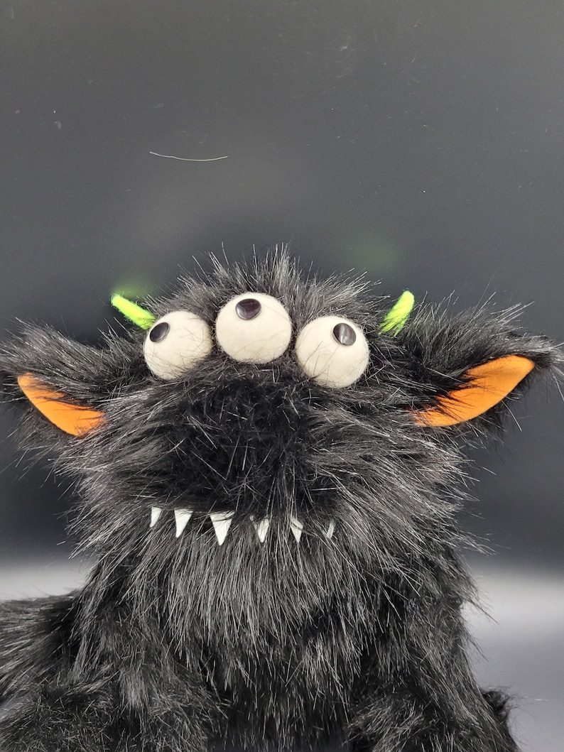 May include: A black furry monster toy with three large white eyes, orange ears, and a toothy grin. The monster has green antennas on top of its head.