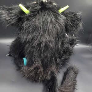 May include: Black plush toy with a long tail and green horns. The toy is sitting on a gray surface.