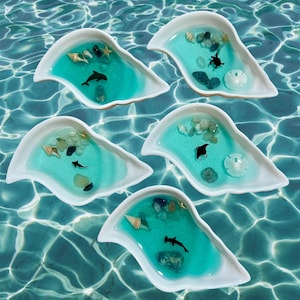 Puede incluir: Cinco platos de cerámica blanca con forma de aleta de delfín, cada uno lleno de resina azul y varias decoraciones con temática marina, incluyendo conchas, estrellas de mar y peces pequeños.