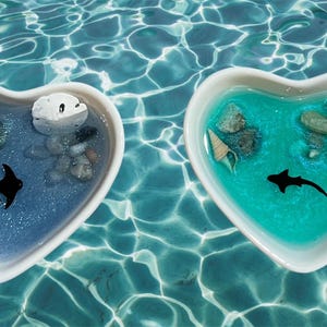 Puede incluir: Dos platos en forma de corazón llenos de resina azul y turquesa, cada uno con conchas marinas, arena y una pequeña silueta negra de un pez. Los platos flotan en una piscina de agua.
